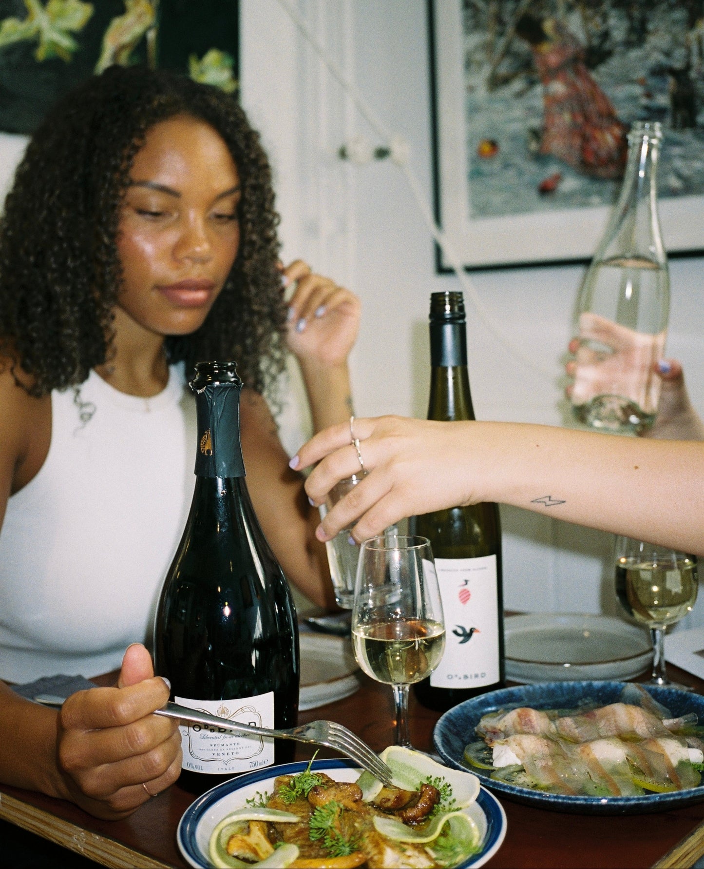 Two women sitting at a table with wine bottles and plates of food, one pouring wine.